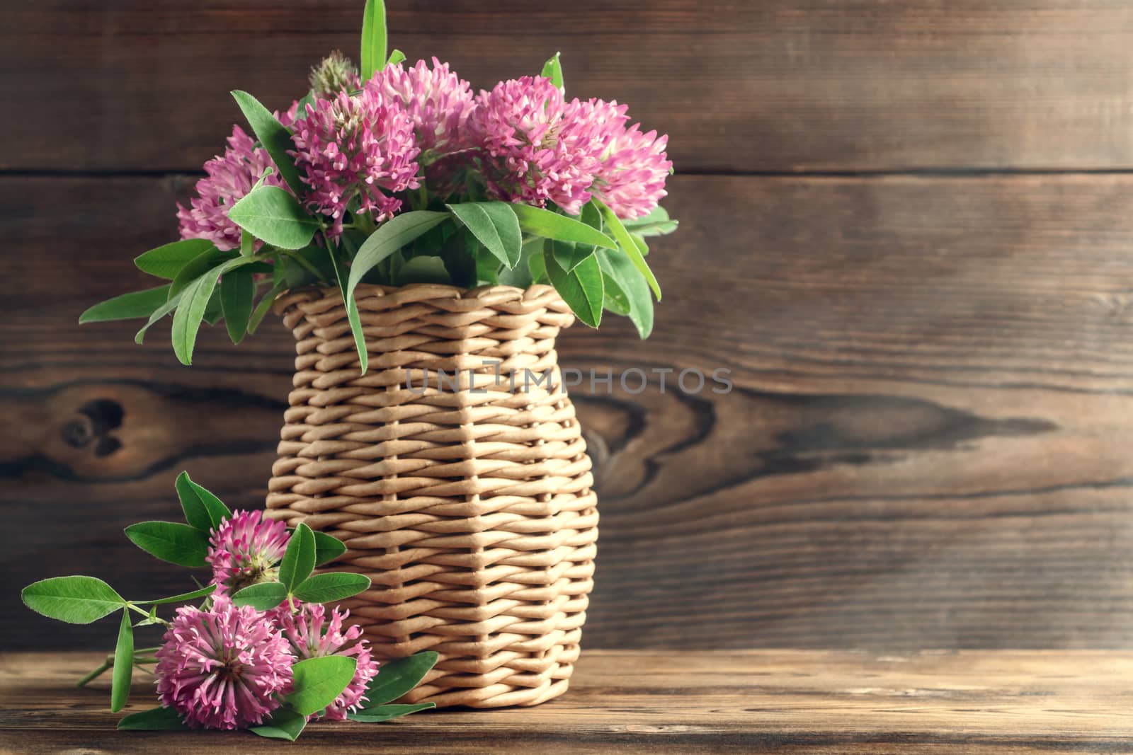 Bouquet of pink clover in a wicker vase on a wooden table on a summer morning.