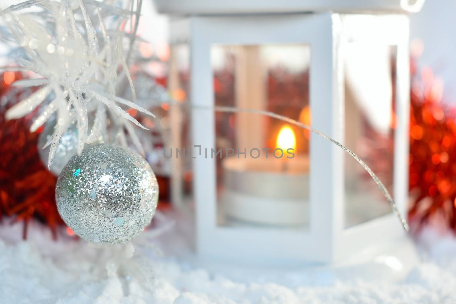 Festive background with red, glittery silver christmas decoration. White lantern on the snow with candlelight. Shallow depth of field. Defocused light, bokeh effect.