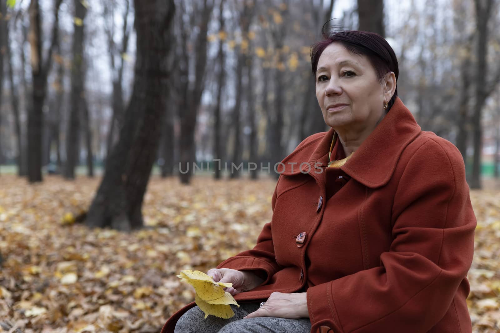An elderly woman in a bright terracotta coat sits thoughtfully in an autumn Park