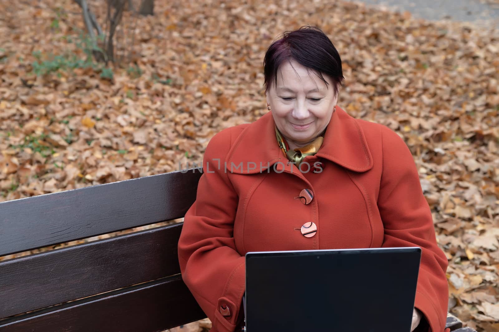 An elderly woman in a bright terracotta coat sits thoughtfully in an autumn Park