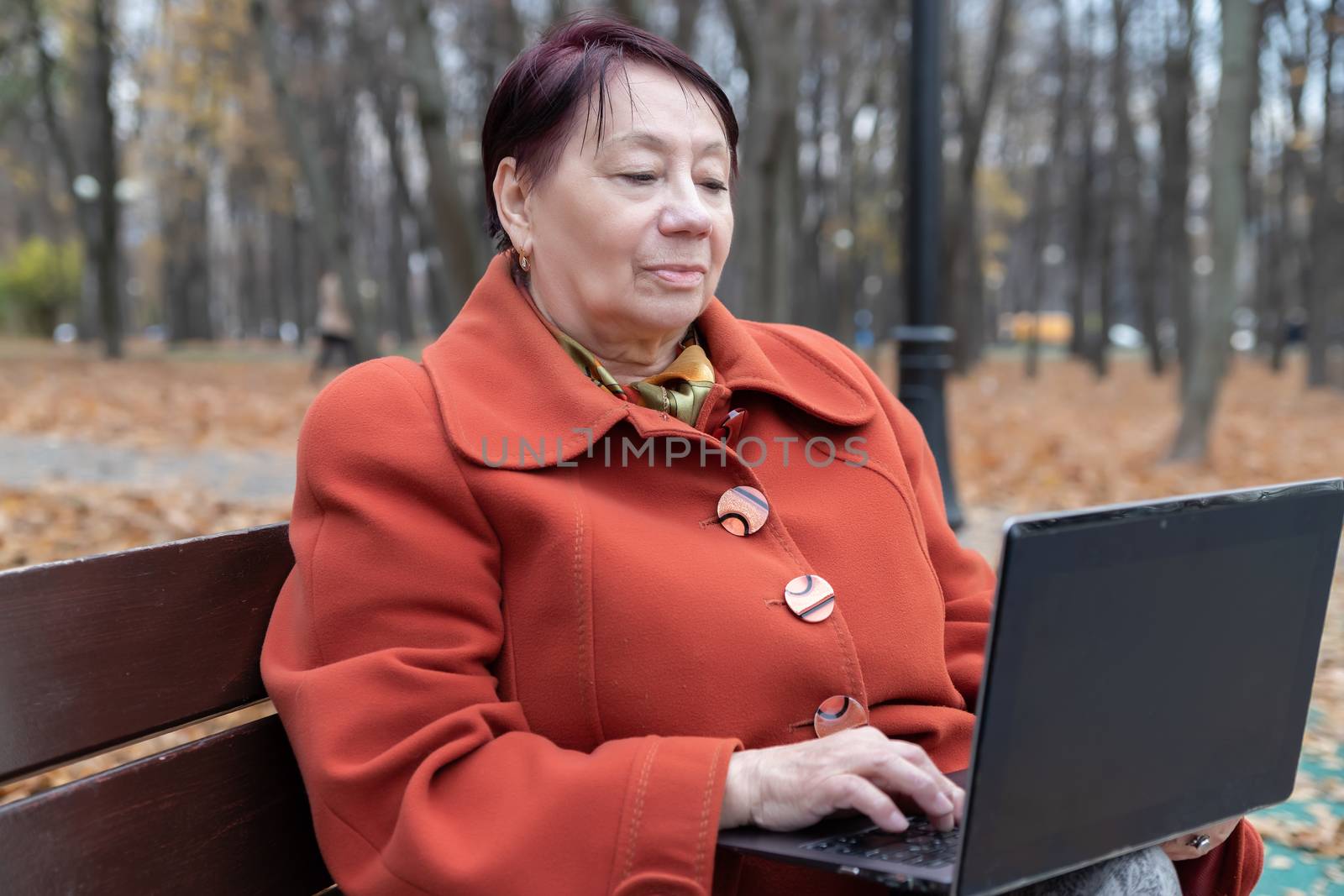 An elderly woman in a bright terracotta coat sits on a bench in an autumn Park with a laptop and selects goods online