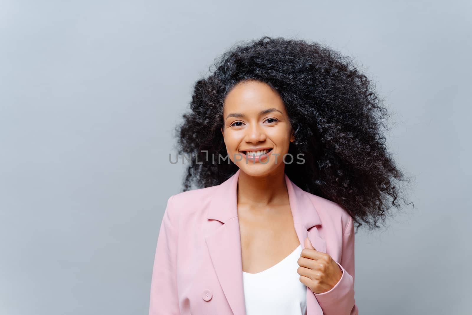 Positive curly dark skinned woman with luminous hair, wears formal purple jacket, poses against grey background with blank space, happy to meet with business partners. Emotions. Happy director by vkstock