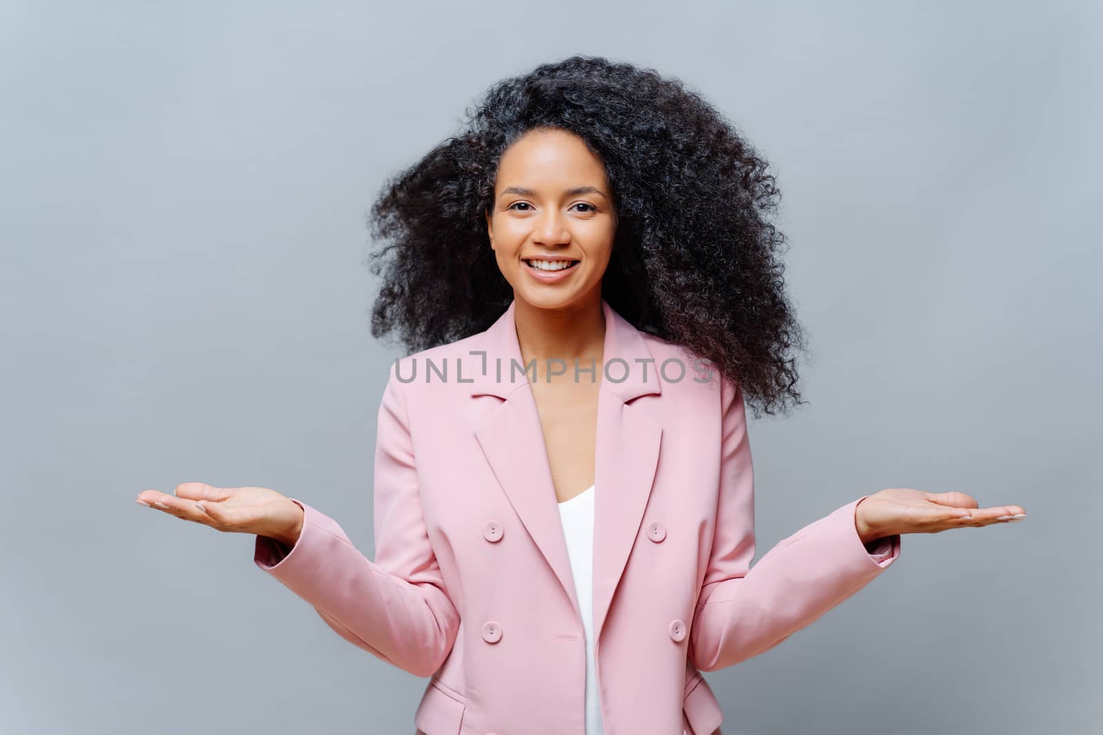 Isolated shot of cheerful female business worker with Afro hairstyle, wears elegant formal violet jacket, raises both palms, presents some product, smiles pleasantly, isolated over grey background.