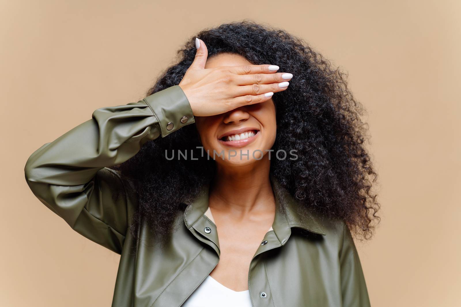 Shy cheerful young Afro woman covers eyes with palm, has toothy smile, hides face, has curly hairstyle, dressed in stylish clothes, isolated over brown background, waits for surprise or gift by vkstock