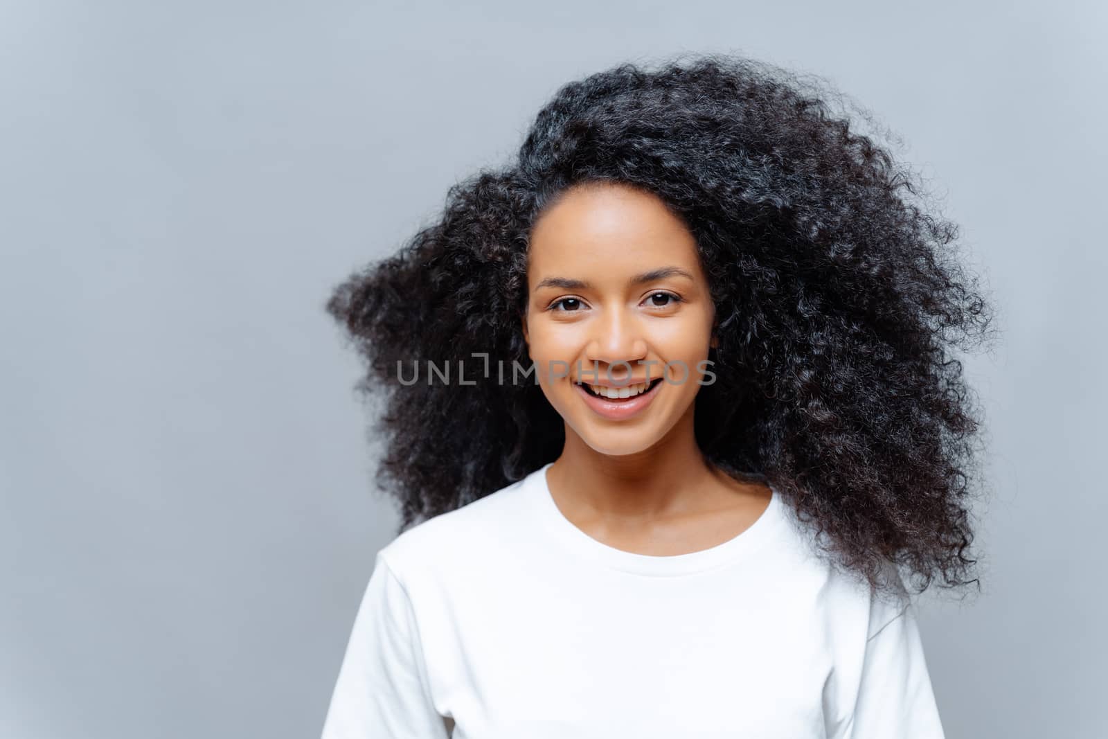 Positive curly woman with natural beauty, dressed in white casual t shirt, has happy expression, looks directly at camera, poses against grey background. Cheerful teenage girl expresses good emotions by vkstock
