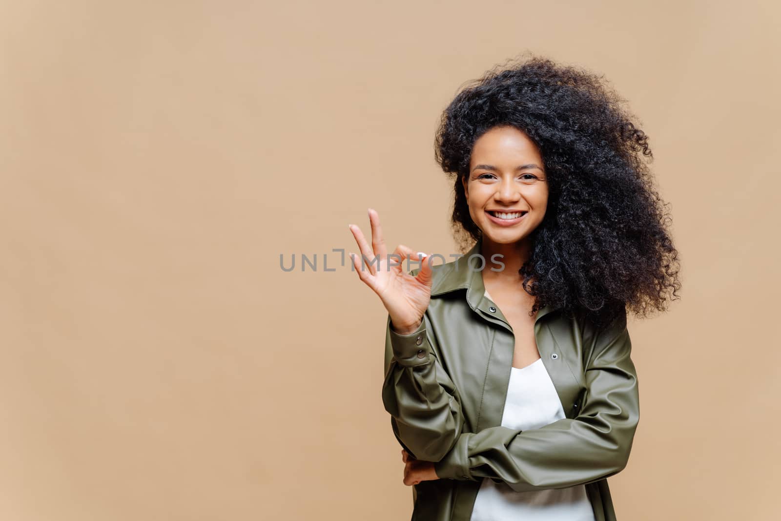 Indoor shot of pleasant looking curly woman has pleasant smile, makes okay gesture, excellent sign, gives approval, dressed in fashionable leather shirt, isolated over brown wall, blank space on left by vkstock