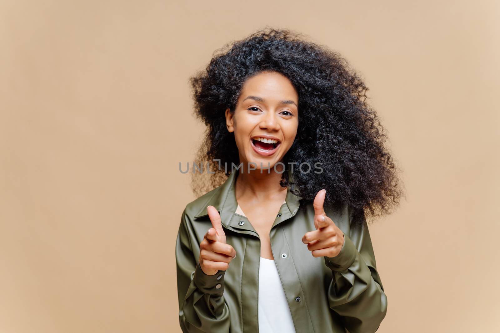 I choose you and order. Positive African American woman points at you, makes finger gun gesture, has overjoyed face expression, dressed in fashionable leather shirt, poses indoor over brown wall by vkstock