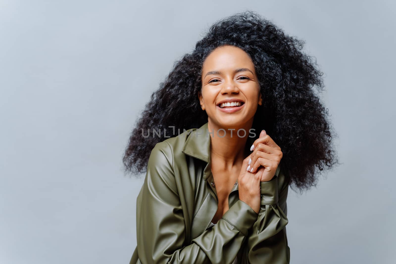 Joyful woman with dark skin, Afro hairstyle, keeps hands together, smiles sincerely at camera, shows white teeth, wears leather shirt, poses over grey background. People, emotions, ethnicity by vkstock