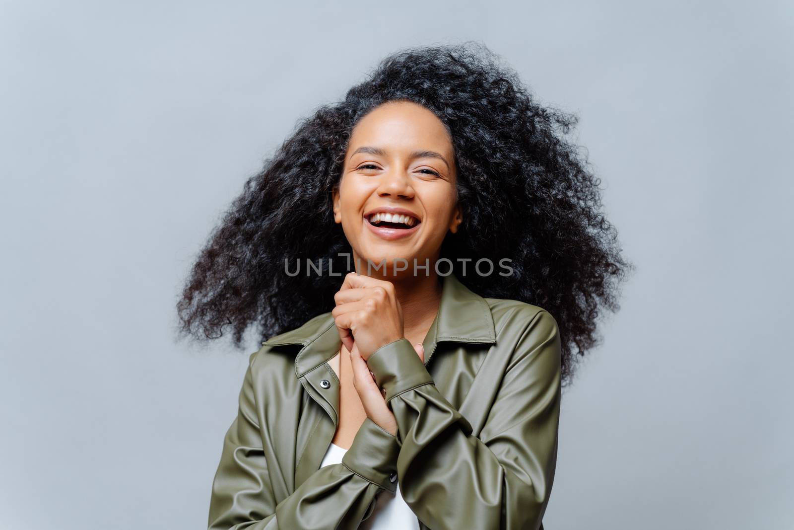 Overjoyed dark skinned curly woman laughs happily, laughs at funny joke, keeps hands pressed together, dressed in fashionable clothes, isolated over grey background. People and positiveness. by vkstock