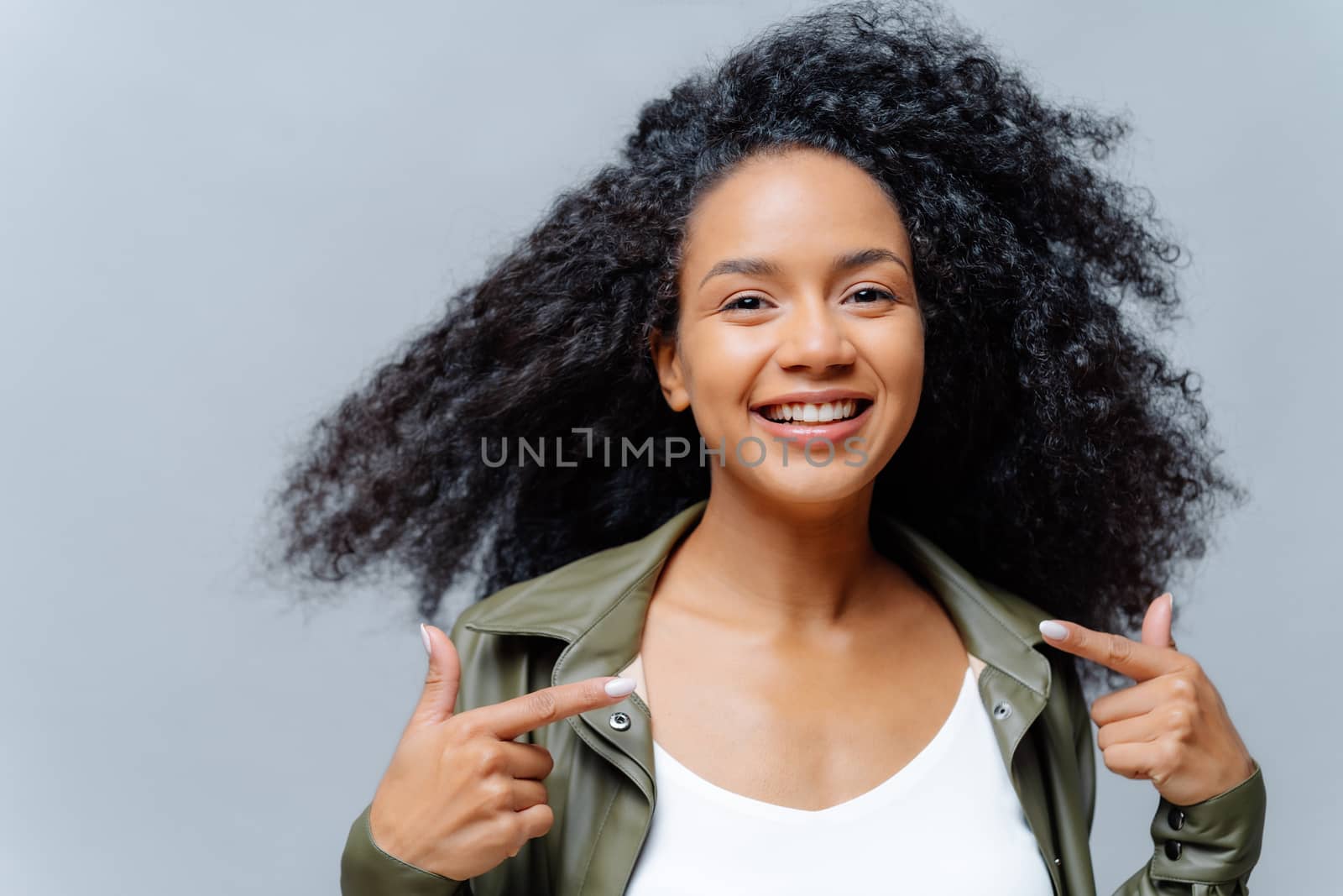 Headshot of pretty smiling woman with Afro hairstyle, points both index fingers at herself, feels proud of herself, wears leather shirt, isolated over studio background. Look at me, my new outfit by vkstock