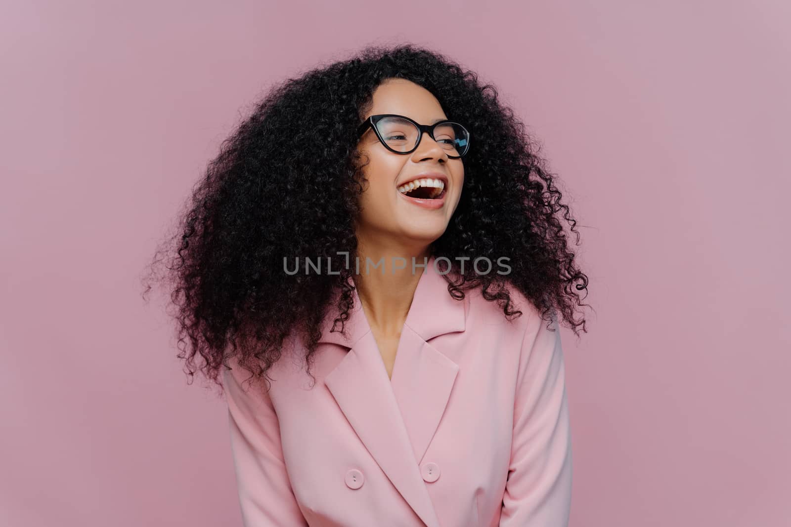 Joyous overjoyed dark skinned female manager has frizzy curly hair, laughs with happiness, wears elegant costume, transparent glasses, focused away, poses over purple background. Emotions concept
