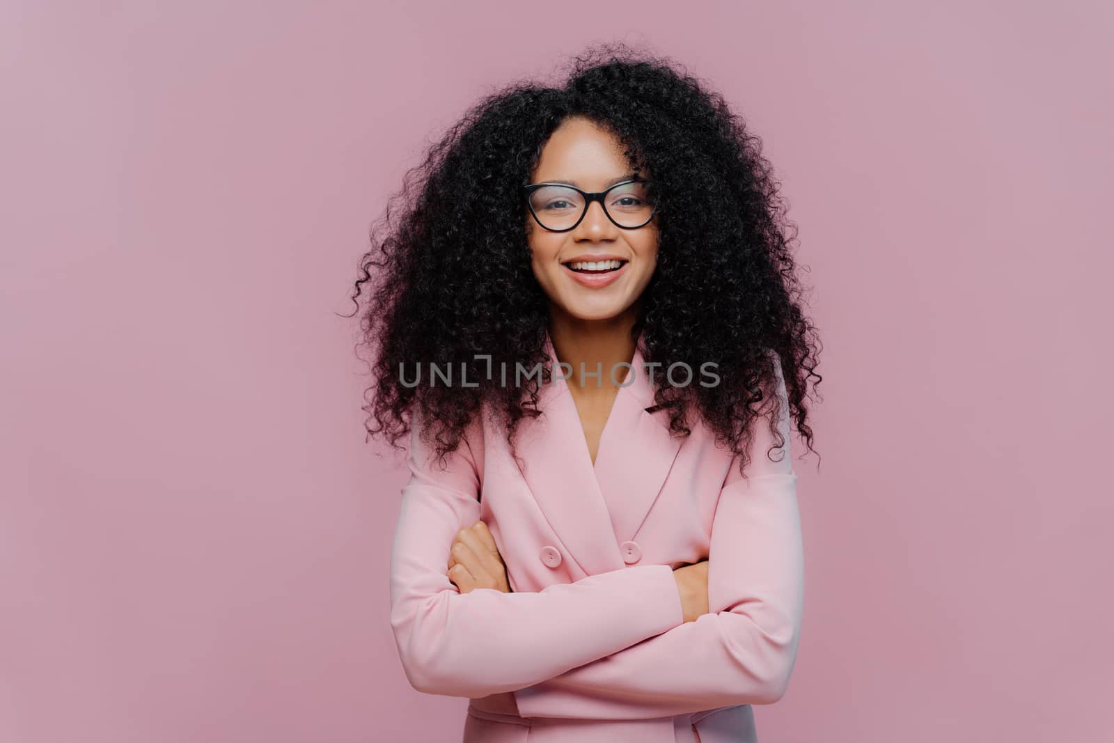 Pleased curly haired woman wears optical glasses for vision correction, elegant suit, keeps hands crossed over chest, poses against purple background, comes on job interview. Businesswoman at work by vkstock