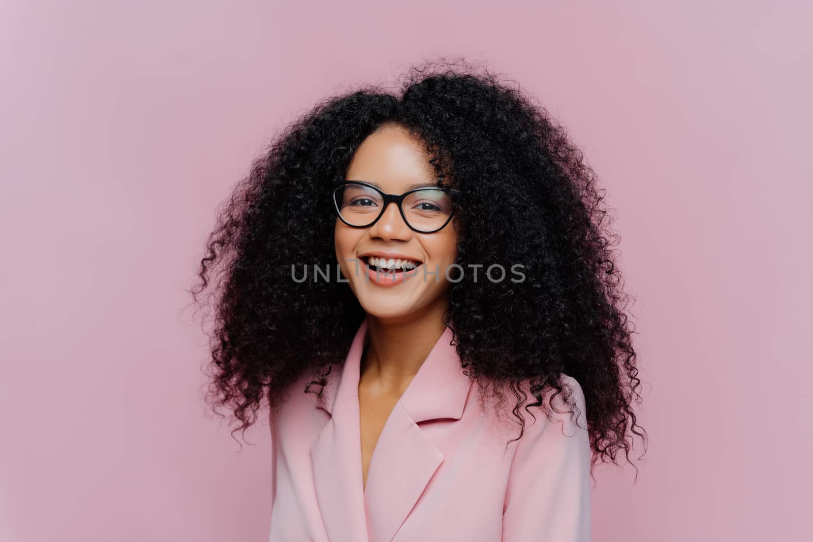 Positive curly haired businesswoman wears spectacles, formal wear, has bushy hairstyle, being in good mood after successful day at work, poses against violet background. Monochrome. Happy director by vkstock