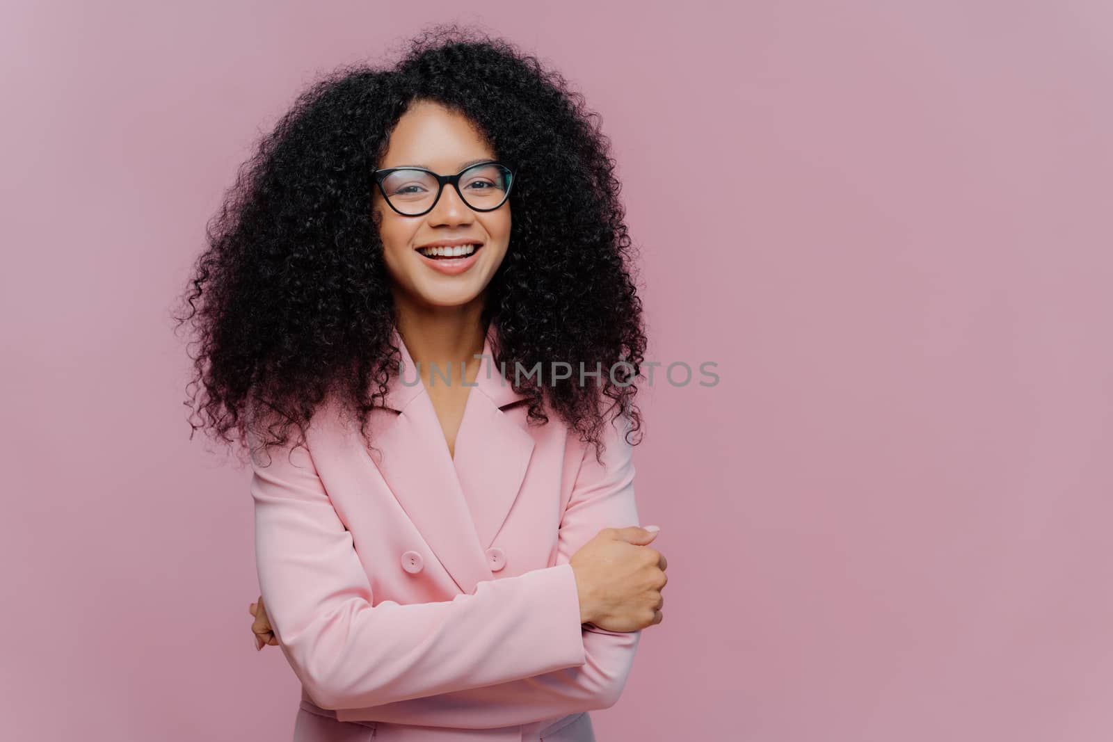 Portrait of happy curly haired businesswoman wears formal jacket, laughs positively, enjoys successful business meeting, has optical glasses, isolated over purple background blank space for your promo by vkstock