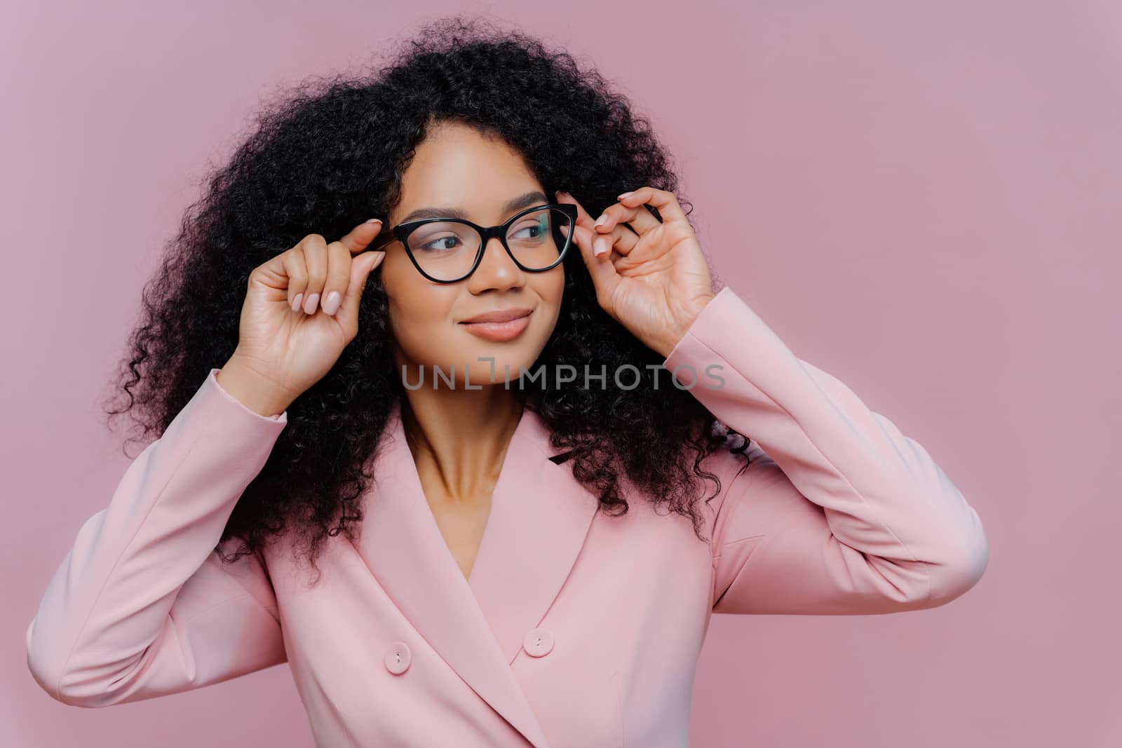 Headshot of attentive female boss keeps hands on frame of glasses, looks pensively away, wears rosy formal suit, has curly Afro hairstyle, poses against purple background. People, business, ethnicty by vkstock