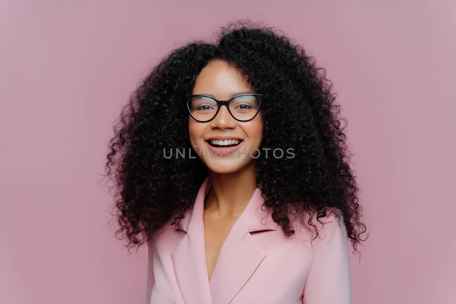 Close up portrait of happy ethnic young woman with crisp hair, looks through optical glasses, wears formal outfit, being in good mood, comes on work, happy get praise from boss, models indoor by vkstock