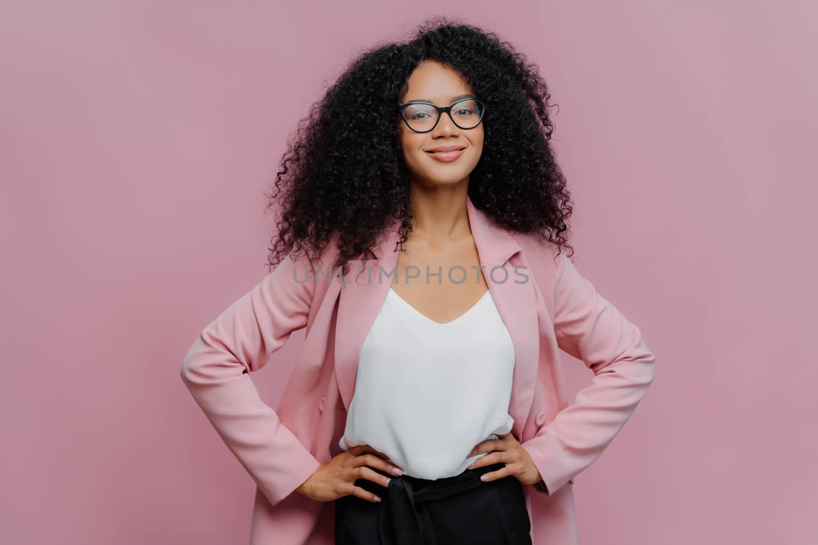 Waist up shot of satisfied dark skinned female office worker keeps hands on her slim figure, has happy look, wears transparent glasses, poses over purple background, ready for talk with colleague by vkstock