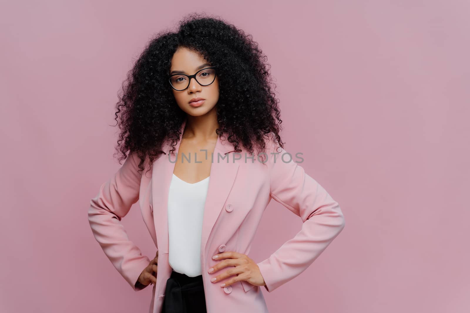 Photo of serious curly haired woman looks confidently at camera, wears formal neat clothes, keeps both hands on waist, wears optical glasses, poses over purple background. People, style concept by vkstock