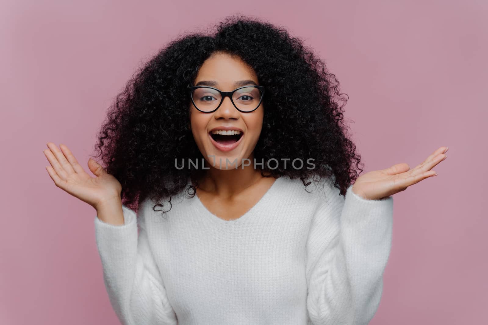 Portrait of joyful curly lady raises arms and spreads palms, wears optical glasses and white sweater, gets pleasant unexpected surprise from close person, isolated on violet studio wall. Happiness by vkstock