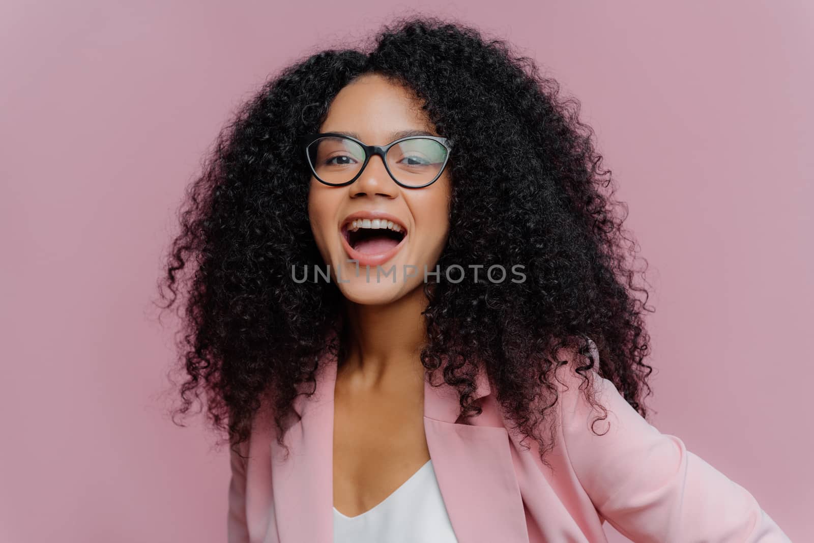 Headshot of overemotive young curly woman keeps mouth opened, wears optical glasses and formal suit, comes on business meeting, has lively conversation with partner, looks gladfully at camera by vkstock