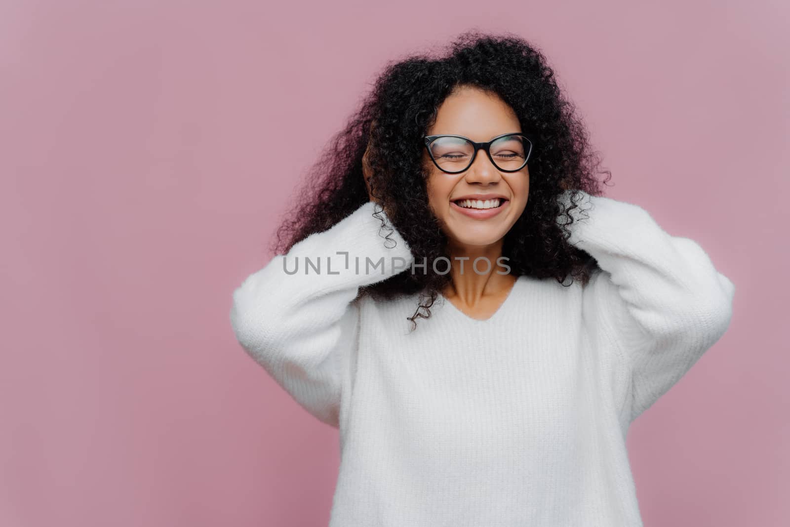 Portrait of joyous pleasant looking African American woman covers both ears with hands, hears loud music, smiles broadly, wears spectacles and white warm sweater, poses indoor, being very emotional