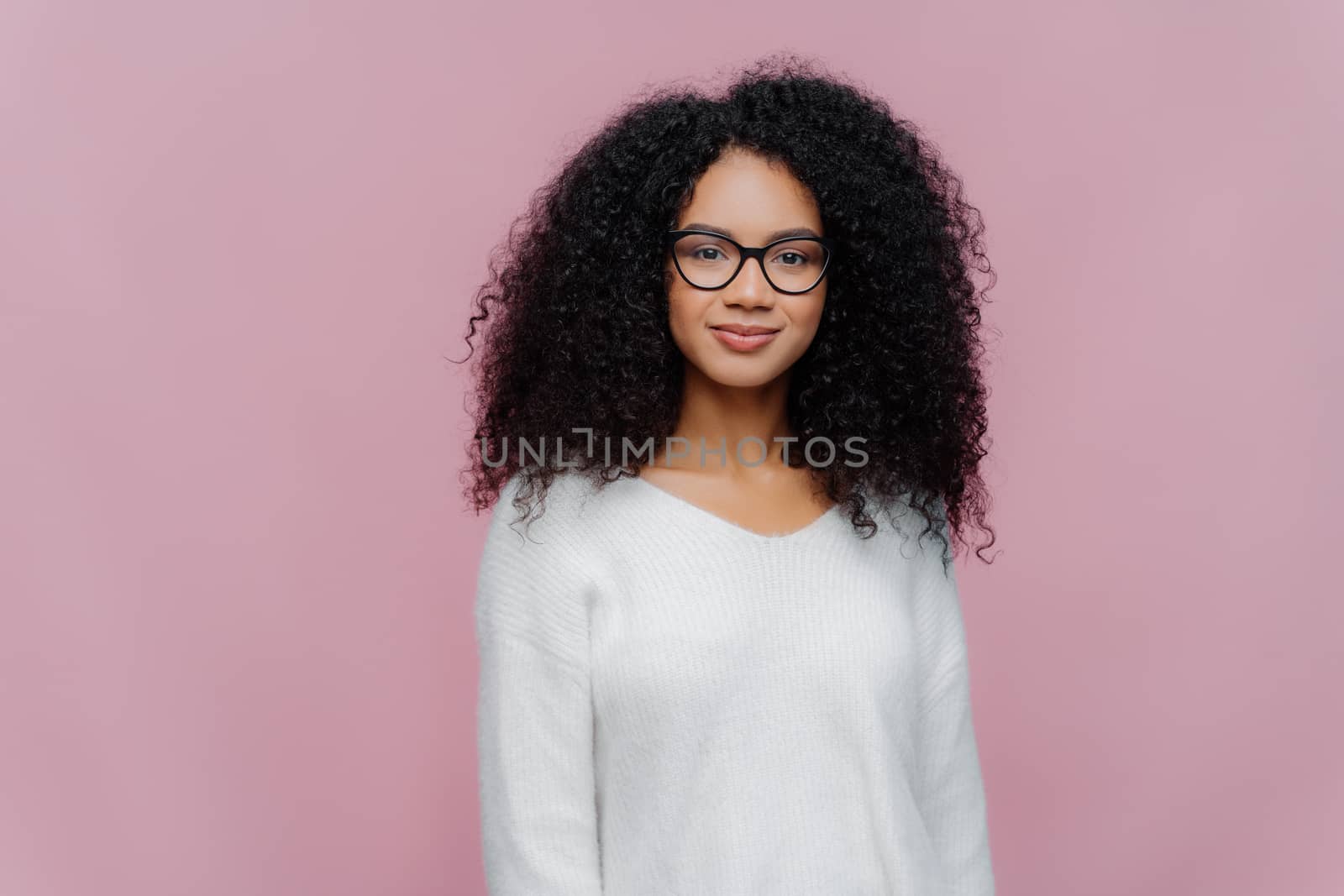 Half length shot of attractive African American woman looks through transparent glasses, white sweater, has serious calm expression, poses against violet studio wall. Facial expressions concept