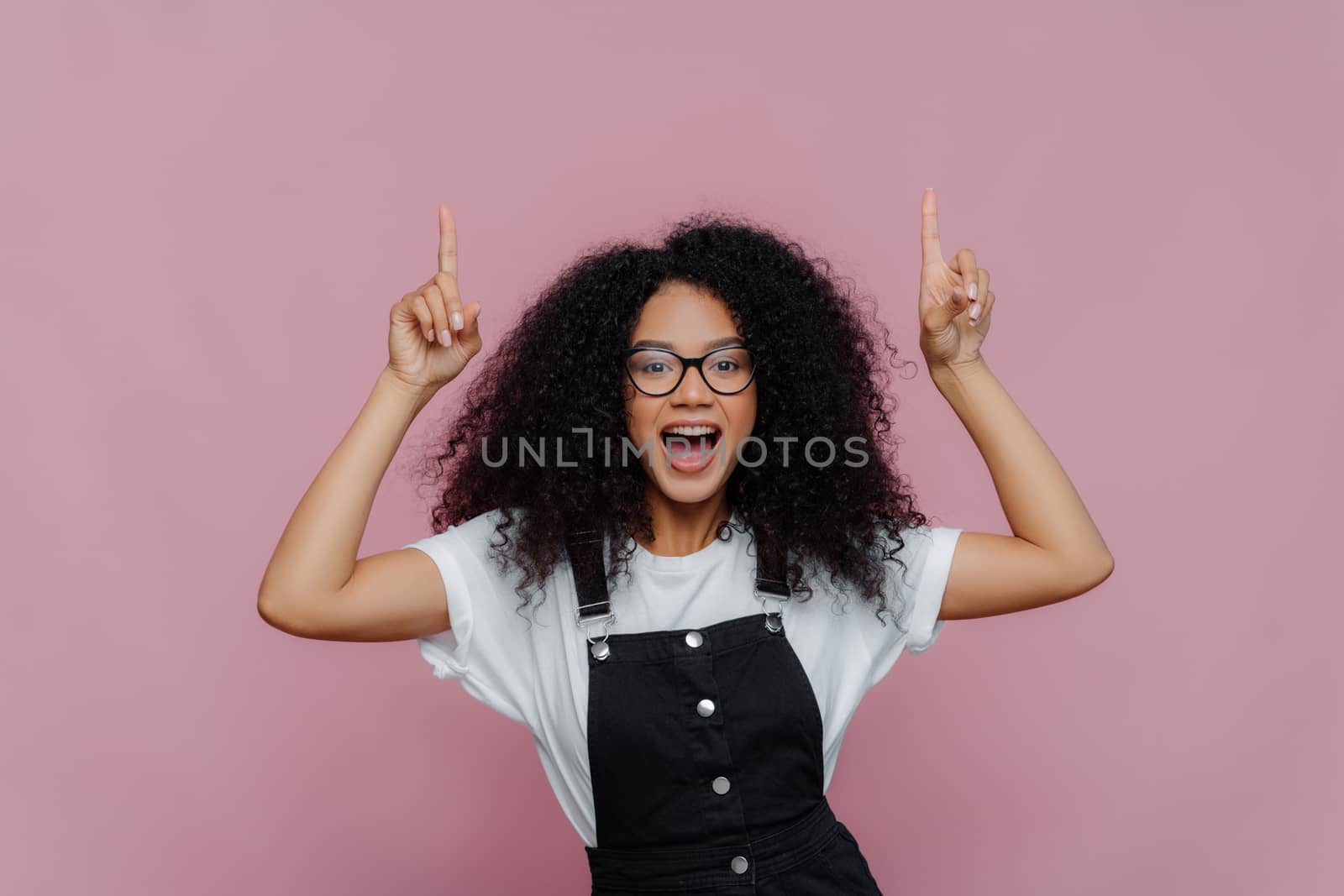 Photo of happy impressed Afro American woman points both index finger above, demonstrates awesome copy space, wears optical glasses and fashionable clothes, poses over violet background. Look above by vkstock