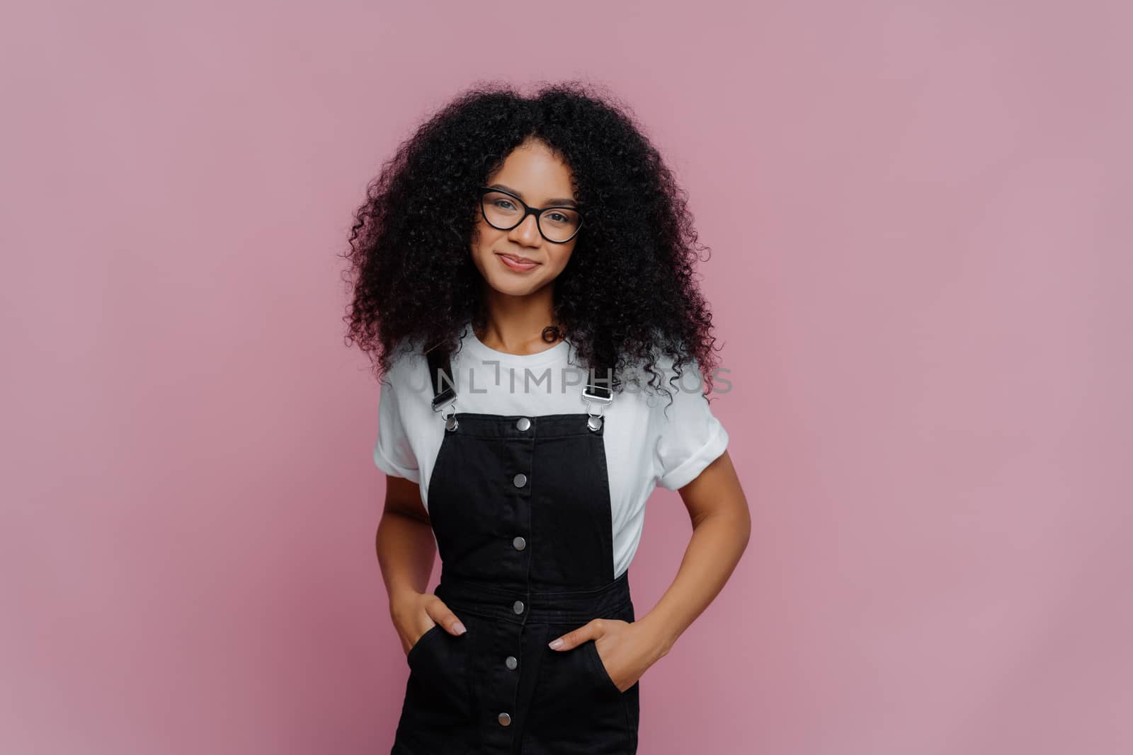 Portrait of happy curly African American woman keeps hands in pockets of sarafan, wears transparent glasses, meets with friends during weekend, models over violet background, has delighted look by vkstock