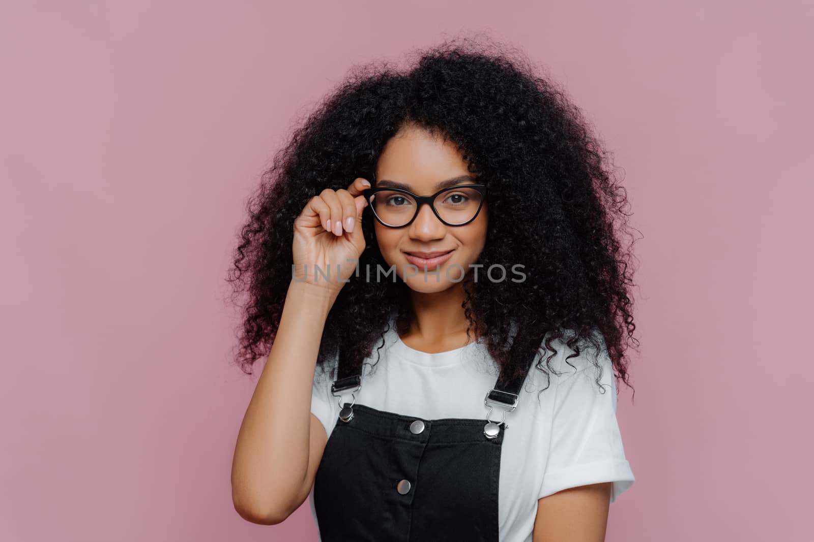 Pretty African American woman keeps hand on frame of glasses, smiles happily, wears white casual t shirt and black overalls, enjoys spare time, poses over violet background. Ethnicity, fashion concept by vkstock
