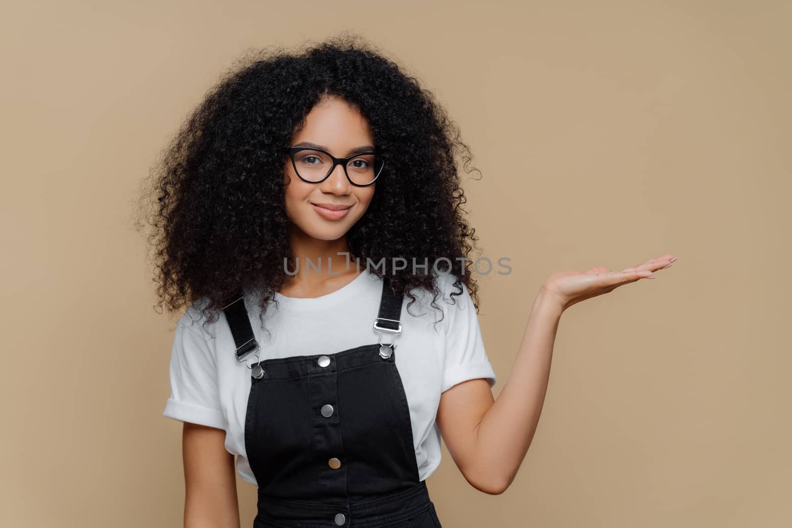 Horizontal shot of pretty dark skinned curly girl raises palm, holds empty space, wears transparent glasses, white t shirt and overalls, isolated over brown background, advertises some object
