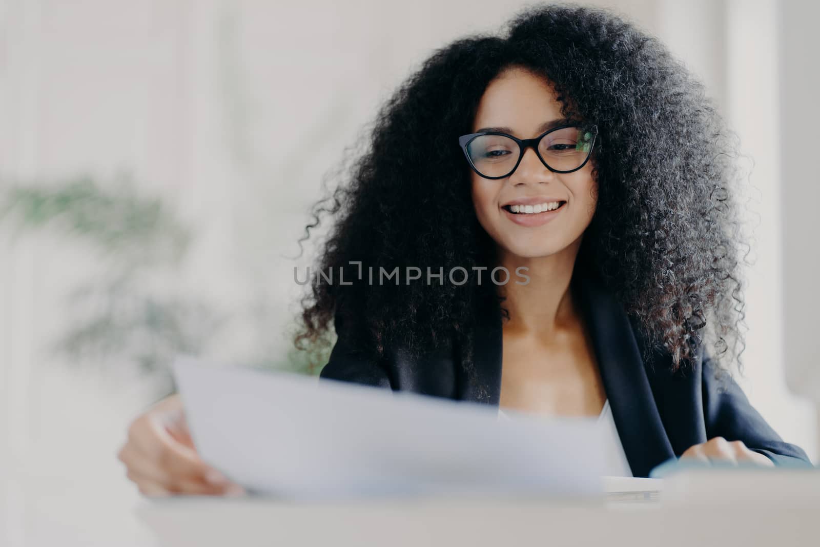 Happy Afro American woman with curly hairstyle, looks through documents, prepares financial report, wears transparent glasses and formal suit, poses in her cabinet, works on financial report by vkstock