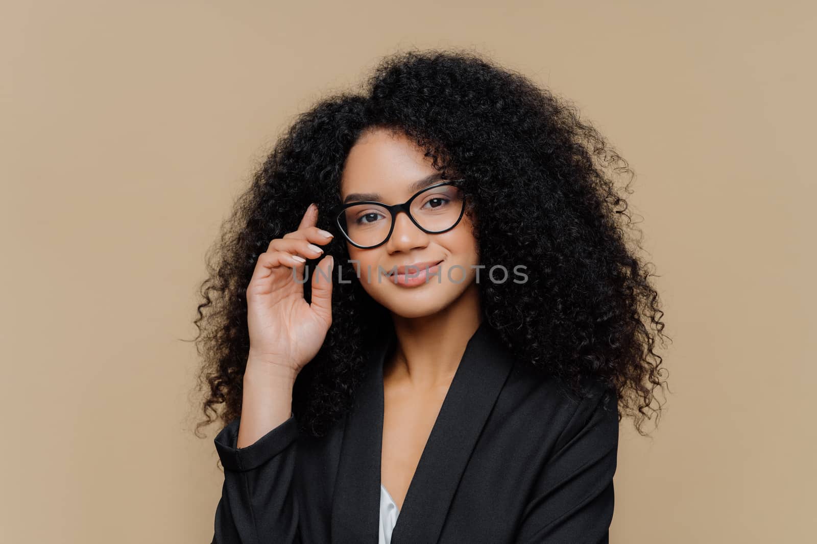 Close up portrait of calm satisfied Afro American woman with bushy curly hairstyle, keeps hand raised, has healthy skin, wears spectacles, black elegant outfit isolated on brown wall. Face expressions