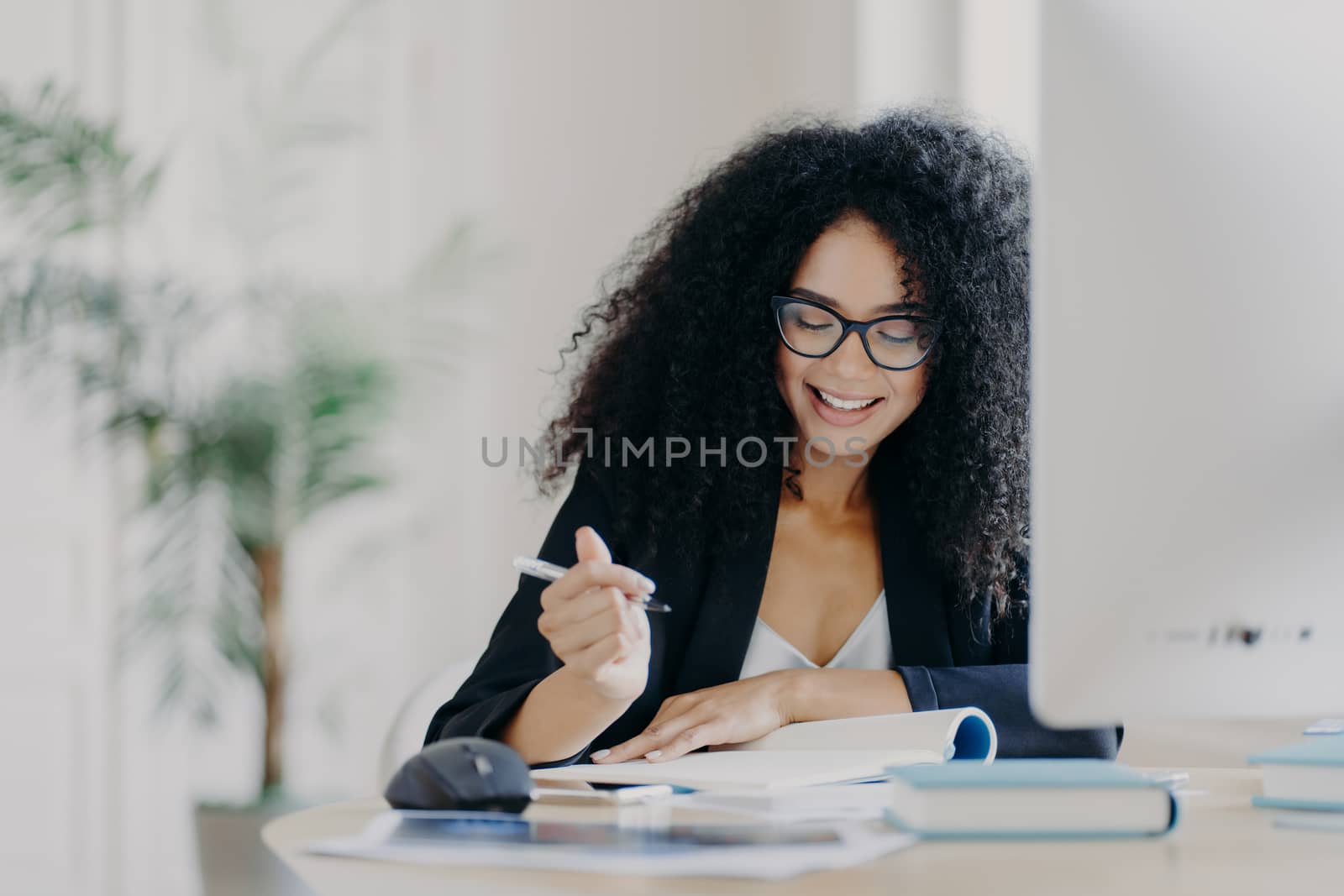 Photo of pleased curly haired woman writes down some information, holds pen, has smile, wears optical glasses and formal clothes poses at workplace with computer. Student writes ideas for course paper by vkstock