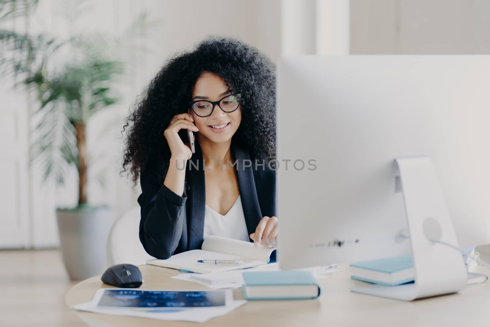 Photo of satisfied Afro American woman calls via smartphone, makes notes in notepad, involved in working process, sits at desk with computer, wears spectacles, formal clothes, has phone communication by vkstock