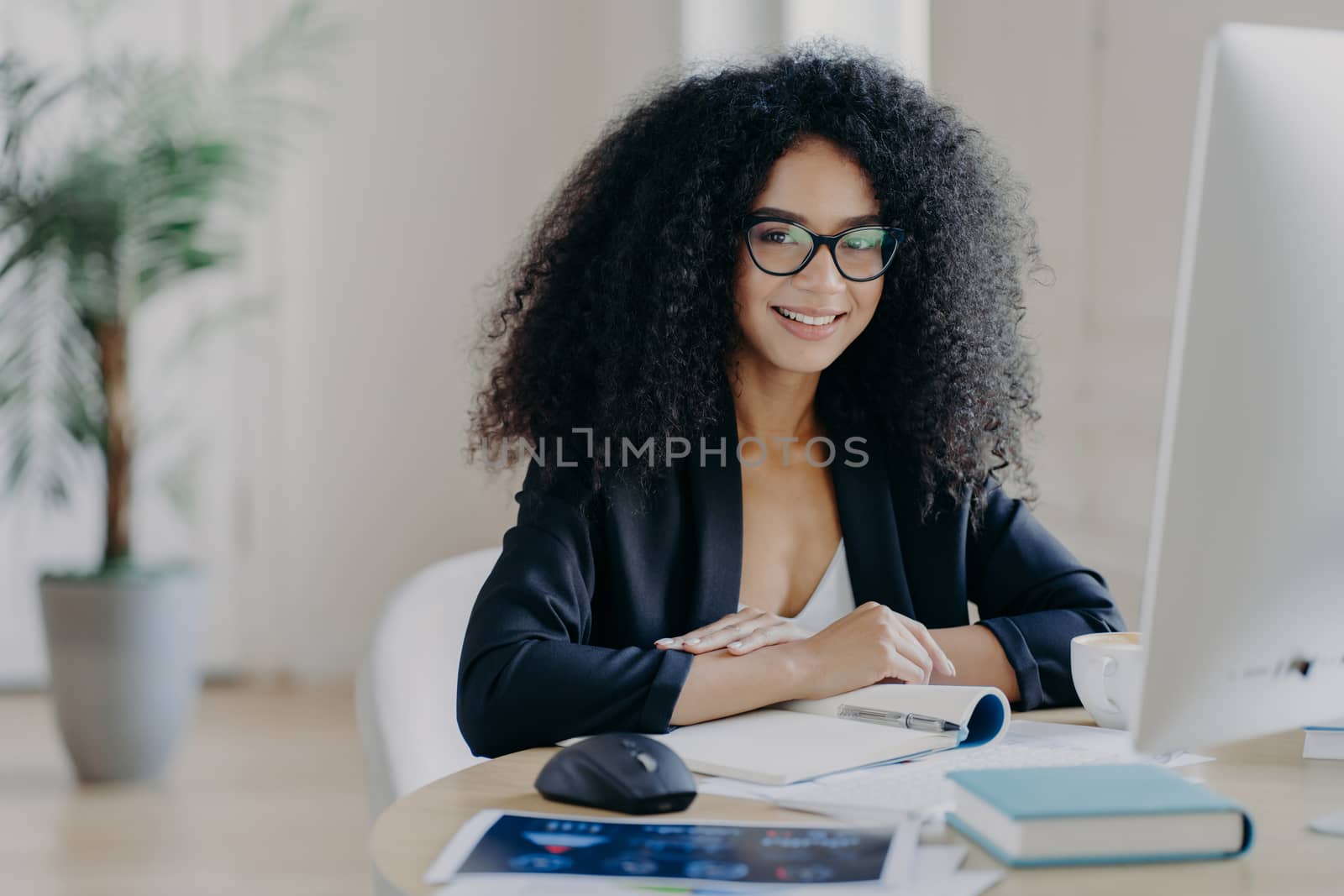 Photo of successful female manager in elegant suit, smiles pleasantly, makes notes, sits at desktop, drinks coffee, poses at computer, smiles positively, poses in office interior. People and work