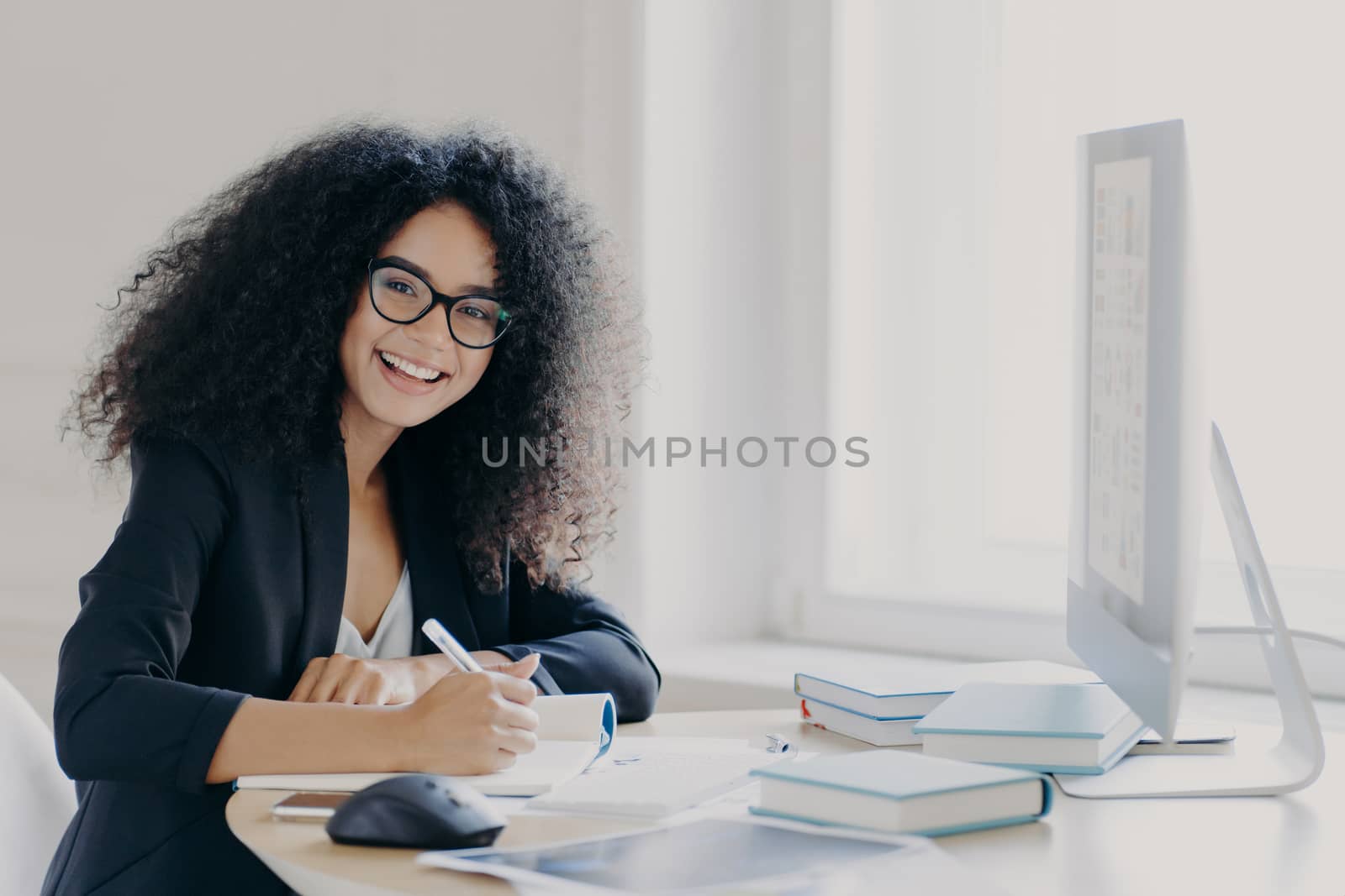 Cheerful female student writes down necessary information in notebook, makes notes, has glad expression, wears transparent glasses and black formal wear, sits at desktop with books and computer
