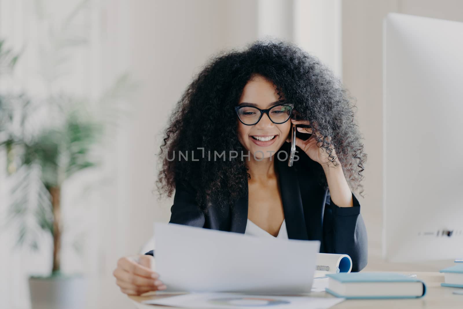 Experienced female CEO has telephone conversation, solves problem, focused in documents, wears spectacles and formal wear, poses at work place, has cheerful expression. Administrative manager by vkstock