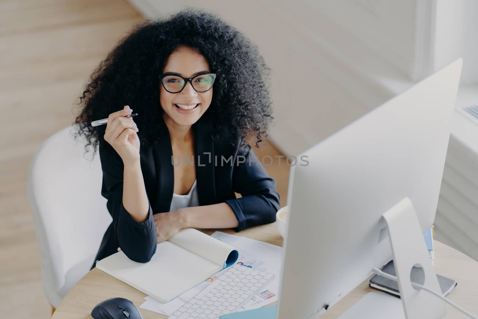 Pleased dark skinned African American woman holds pen, writes down some information, works as editor, looks gladfully at camera, holds pen, uses modern computer for searching something in internet