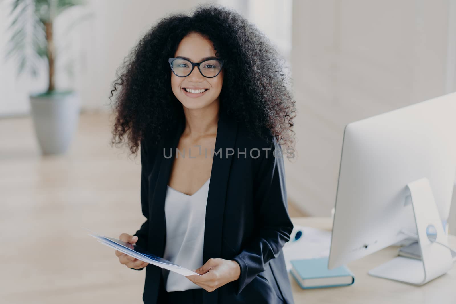 Photo of lovely African American woman holds some papers, being consultant manager, wears elegant clothes and spectacles, stands near desktop with computer and textbooks. Happy businesswoman