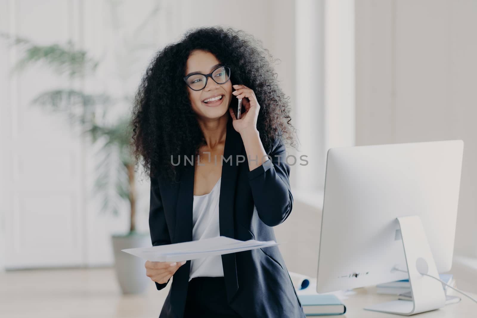 Busy female manager holds mobile phone near ear, holds papers, checks report, has telephone conversation, wears glasses and elegant wear, smiles broadly, workplace with computer in background