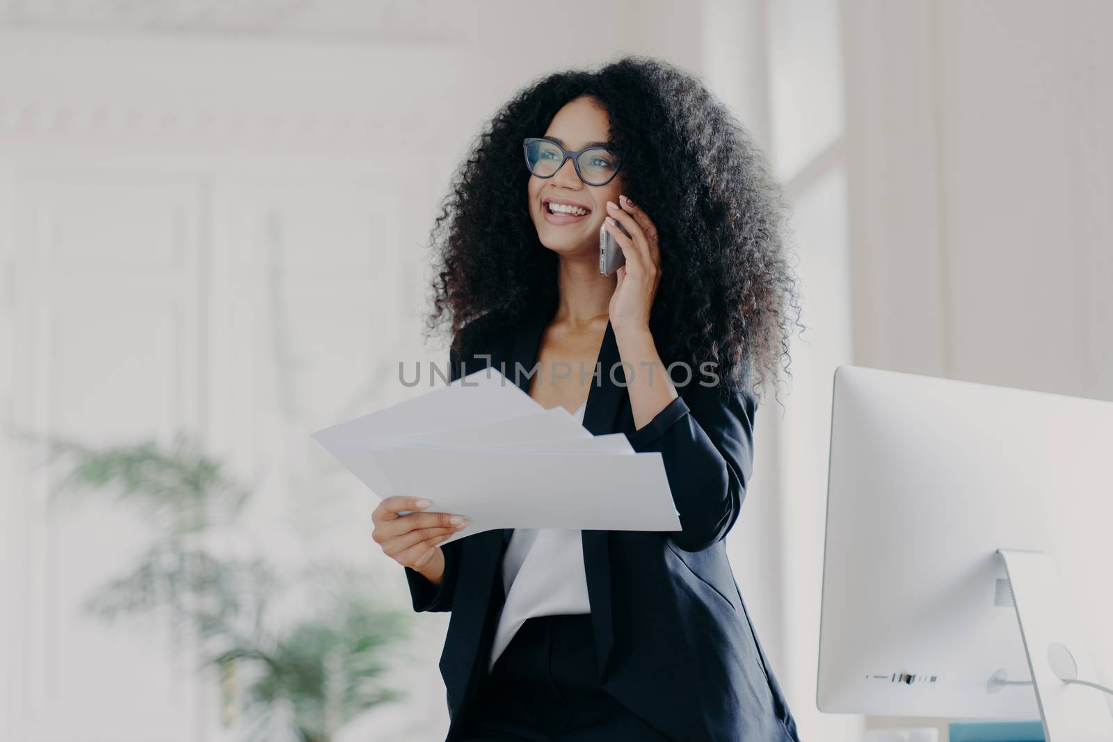 Pleased successful businesswoman with Afro hairstyle wears spectacles to provide eyes protection, holds documents, makes telephone call to business partner, stands in cabinet near workplace.