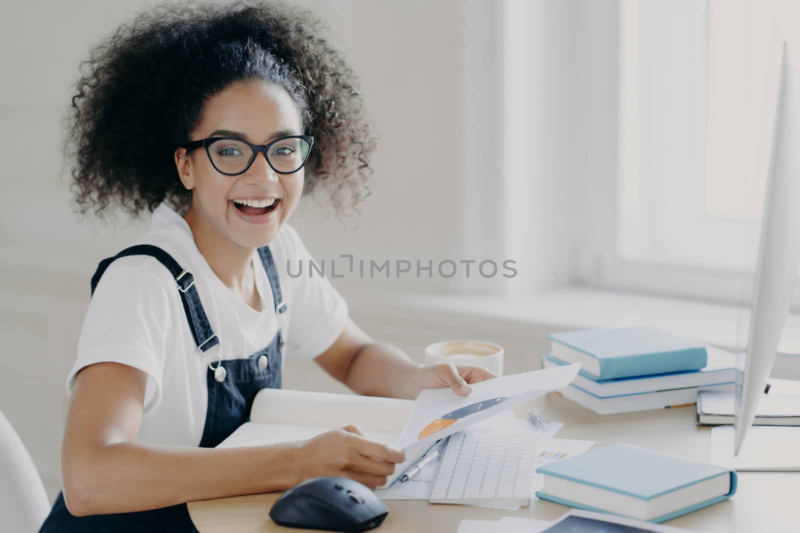 Sideways shot of happy prosperous female entrepreneur holds paper, studies chart, makes planning, being busy with paper work, sits at wooden desk with computer, wears glasses, t shirt and overalls