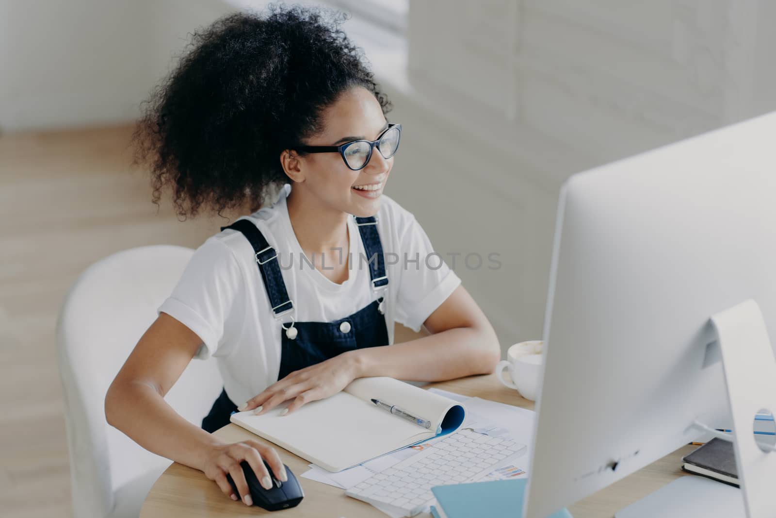 Glad young African American woman sits in front of modern computer, makes project work, has many papers and notepad on table, wears spectacles for vision correction, dressed in casual clothes