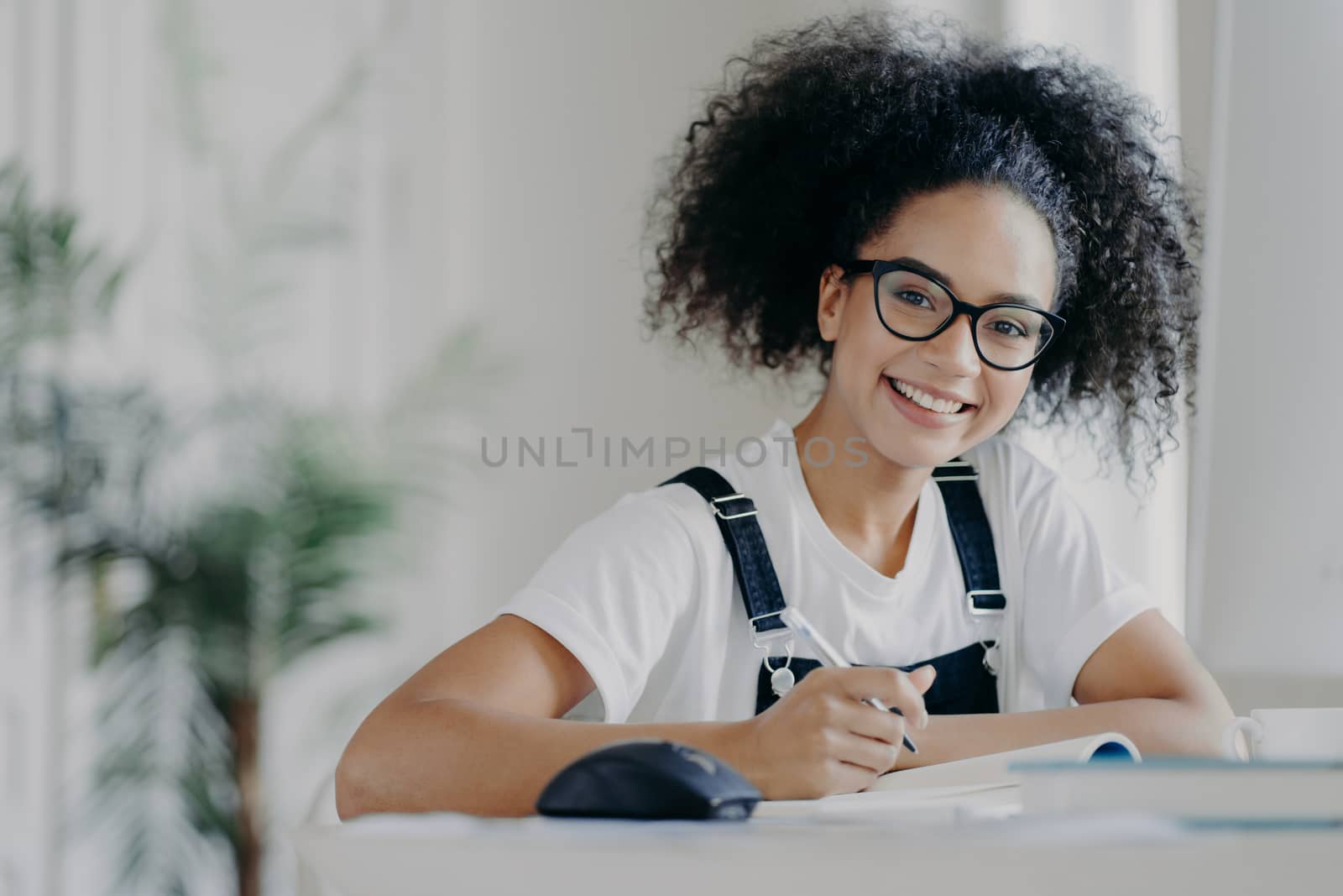 Time for studying. Happy dark skinned teenage girl holds pen, writes down information needed for course work, busy preparing for classes, dressed in casual wear and spectacles, being hard working by vkstock