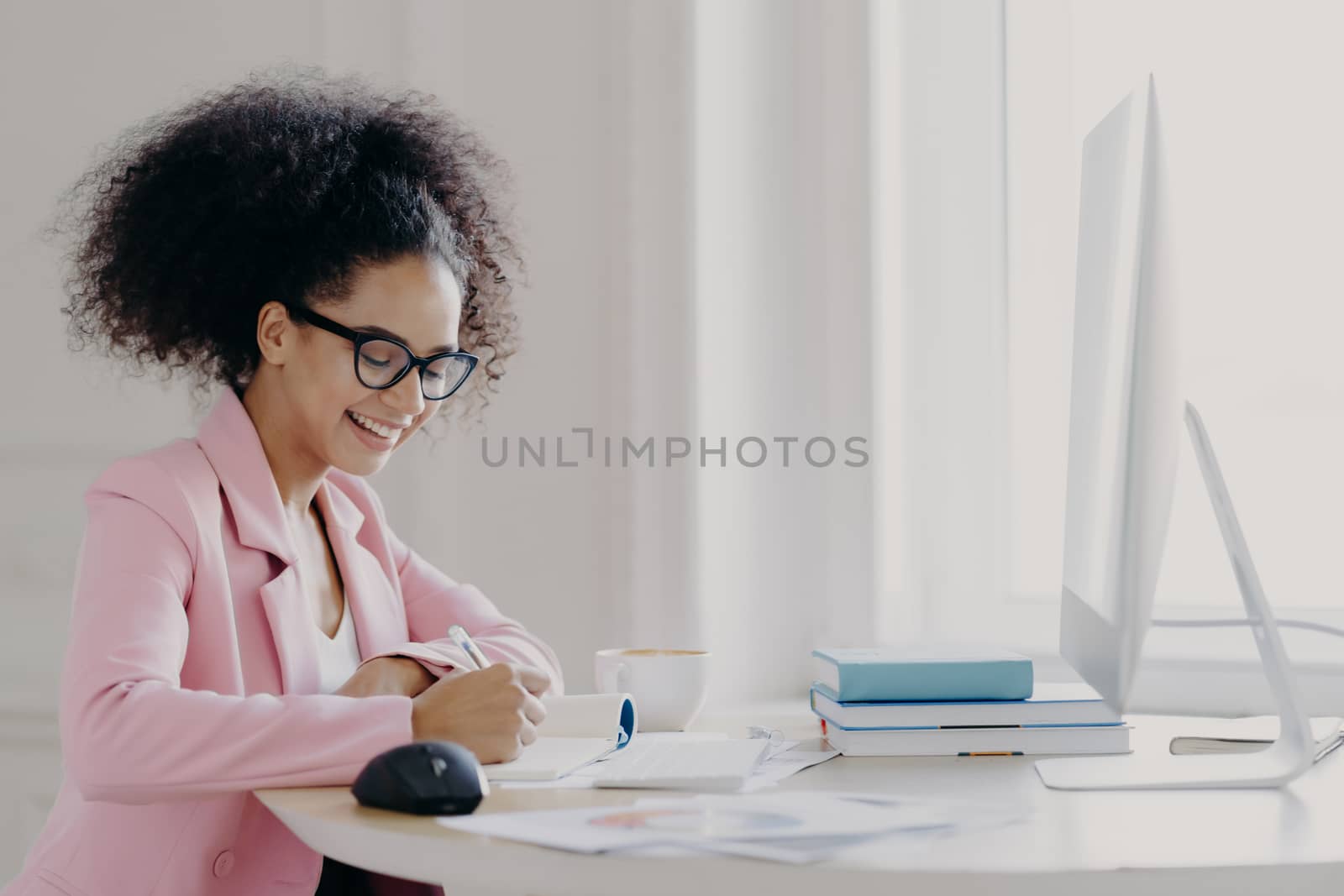 Photo of happy curly businesswoman dressed in elegant rosy suit, writes down something in notepad with cheerful expression, uses computer for searching information online, studies or works indoor