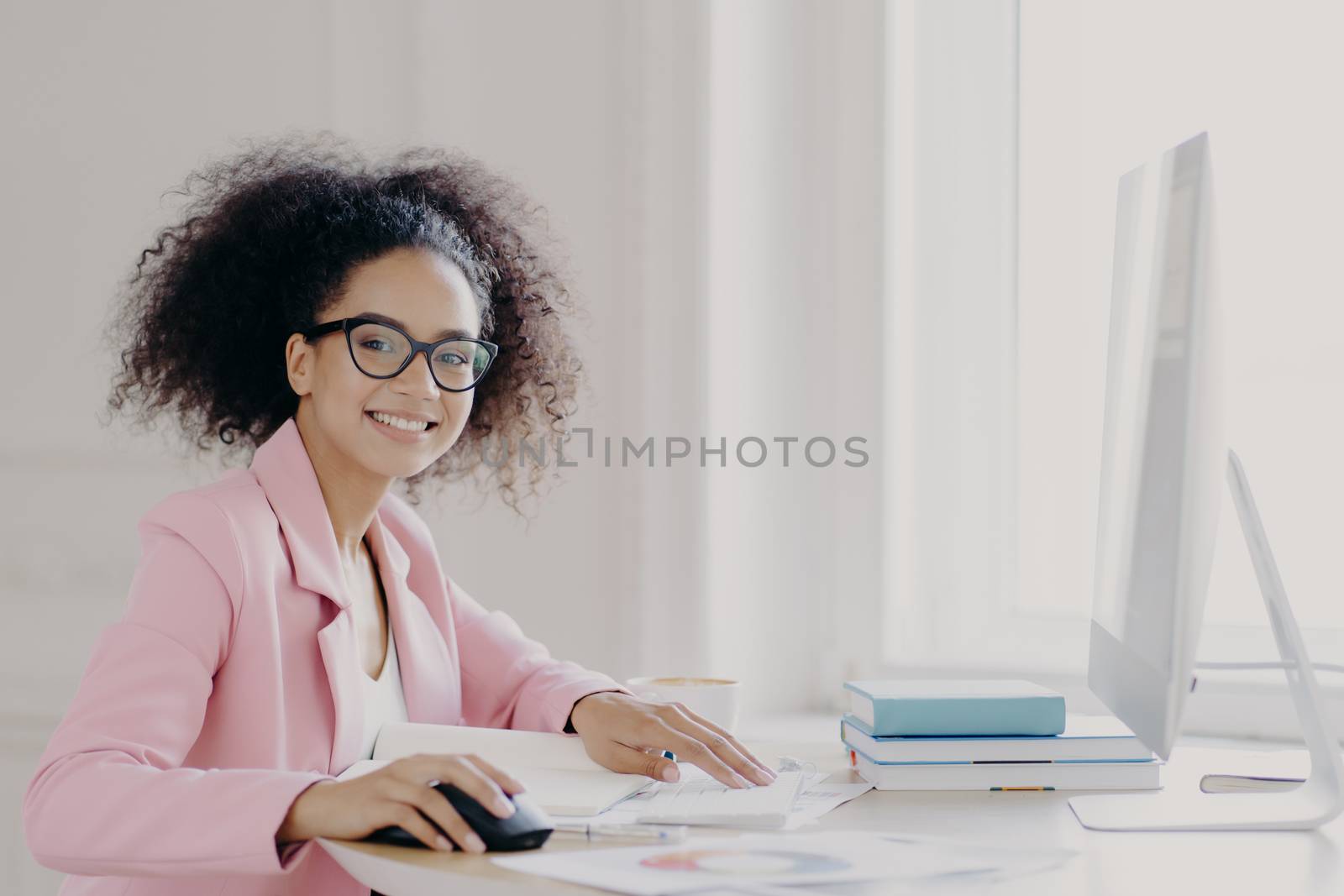 Prosperous curly female entrepreneur checks documents, studies contract, works on computer, wears glasses and pink jacket, poses at workplace, smiles broadly, happy almost finishing work in time