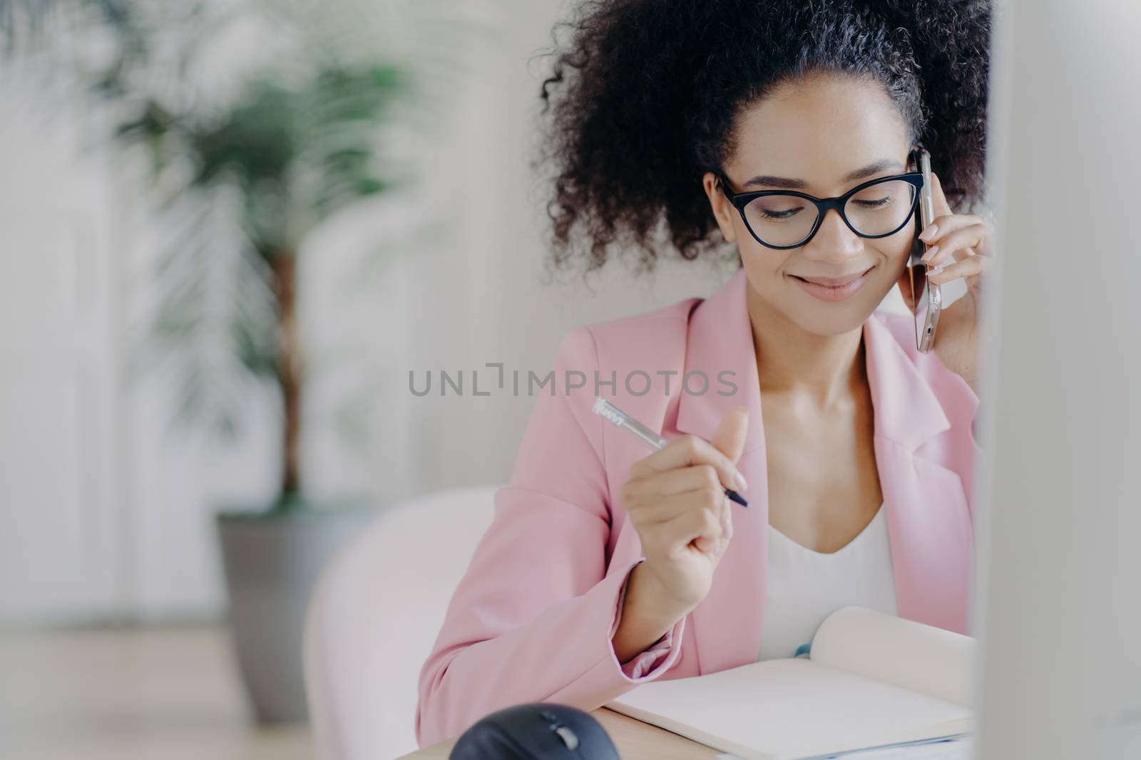 Smiling charming woman with curly hair, writes down information in notebook, gets consultancy via cellphone, wears glasses and elegant suit, busy working, owns business company, solves problems by vkstock