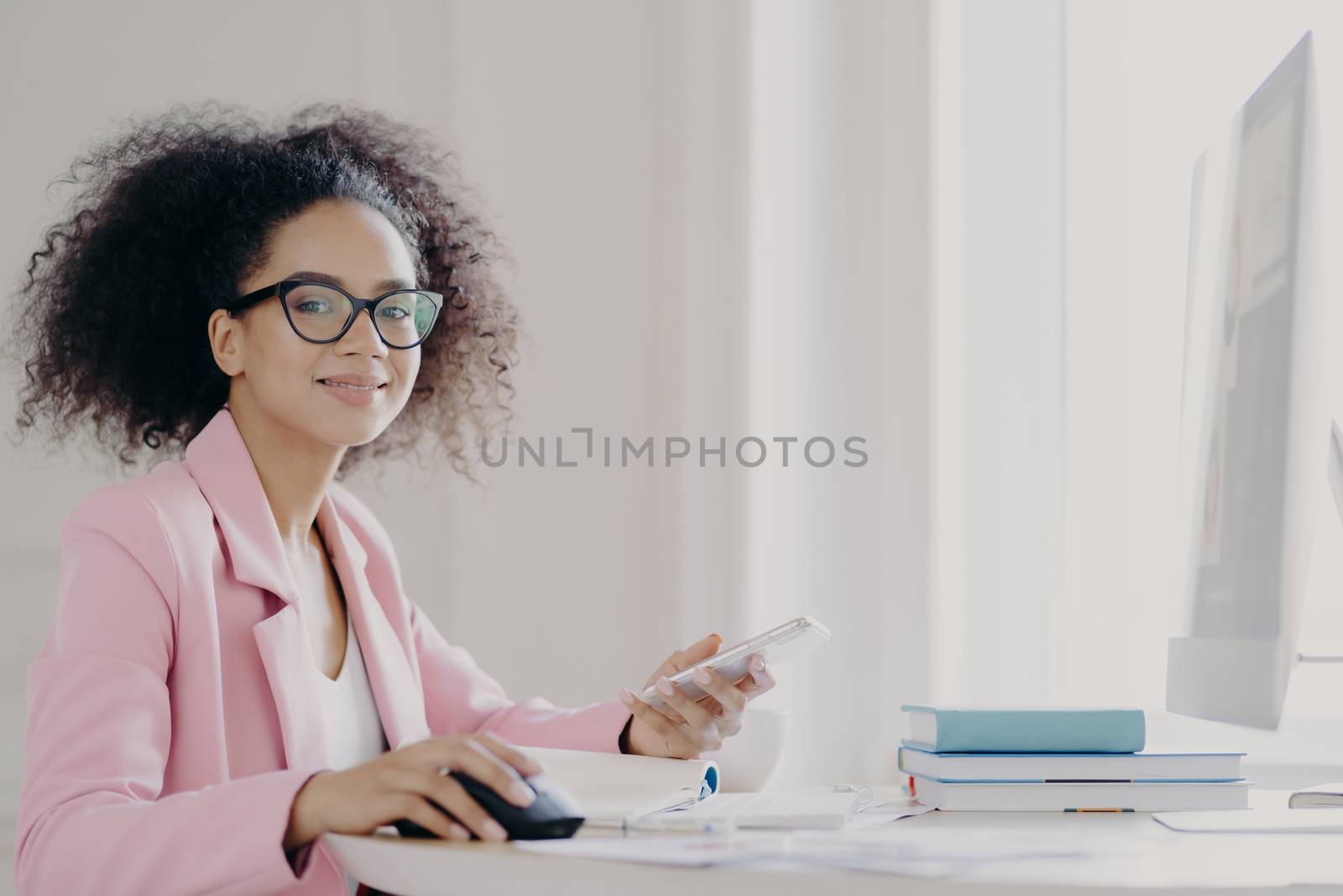 Cheerful dark skinned lady holds mobile phone in hand, waits for call, sits in front of computer, wears optical glasses and elegant suit, poses in her cabinet being busy at work. Technolgy, occupation by vkstock