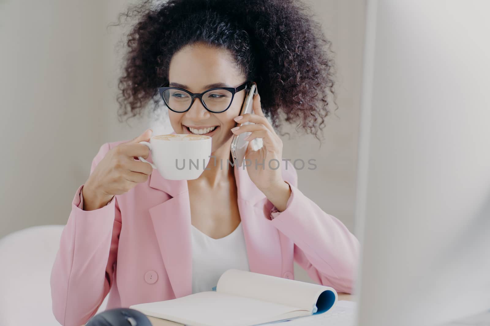 Cropped image of happy young African American woman makes phone call, drinks aromatic latte or espresso, poses in office interior, has pleasant smile wears optical glasses and rosy formal jacket