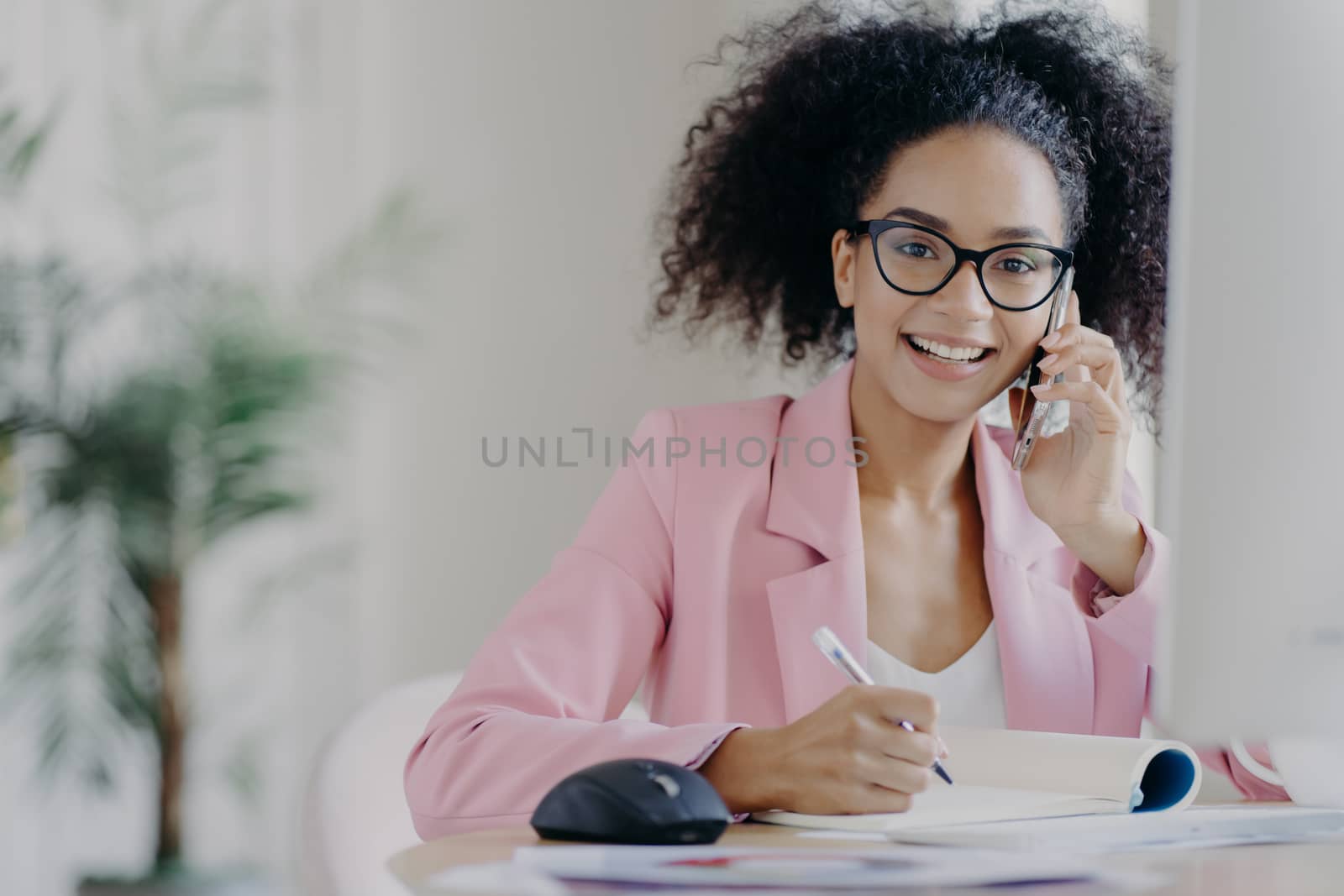 Happy African American woman with curly hairstyle, writes some information, speaks via cell phone, sits at desk against blurred office background. People, technology, communication, business concept